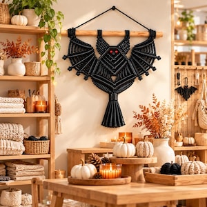 Puede incluir: Un tapiz de macramé negro con forma de murciélago y dos ojos rojos, suspendido de un listón de madera horizontal mediante un cordón negro. El murciélago presenta intrincados patrones de nudos que forman las alas y la cola.