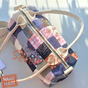 Może przedstawiać: Cylindryczna torba podróżna typu patchwork wykonana z różnych kwadratów materiału w dżinsowe, różowe wzory w kratkę gingham oraz motywy kwiatowe. Torba posiada złoty zamek błyskawiczny, kremowe paski z taśmy oraz znak wodny Seven Sisters.