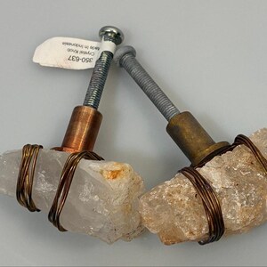 May include: Two cabinet knobs featuring raw, translucent white crystal stones wrapped in dark copper wire. Each knob has a threaded metal bolt extending from a cylindrical base, one in copper and one in brass, with a small white tag attached.