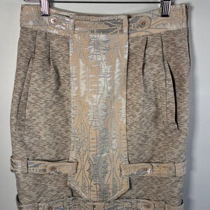 May include: A knee-length skirt featuring a heathered brown fabric base with a central vertical panel and horizontal straps in a metallic silver and beige patterned jacquard. The skirt includes a high waistband, side pockets, and button details.