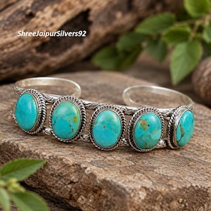 May include: A silver cuff bracelet featuring five oval-shaped turquoise stones set in twisted rope-style bezels. The metal band displays an engraved pattern along the sides. The text ShreeJaipurSilvers92 is visible in the upper left corner.