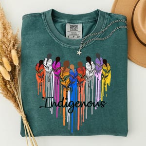 May include: A dark teal crewneck t-shirt featuring a graphic of multiple colorful feathers arranged in a heart shape with the word "Indigenous" written in black cursive script below. The shirt has a Comfort Colors brand label at the neck.