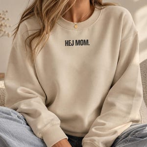 May include: A beige crewneck sweatshirt featuring the text HEJ MOM. printed in black on the upper chest. The garment has long sleeves with ribbed cuffs and a ribbed neckline, paired with light blue denim jeans and a gold chain necklace.