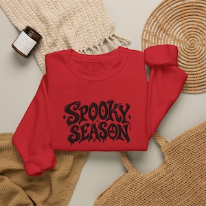 May include: A red long-sleeve crewneck sweatshirt featuring the black text "SPOOKY SEASON" in a stylized, dripping font with small star accents. The letter "O" in "SEASON" is replaced by a jack-o'-lantern face graphic.