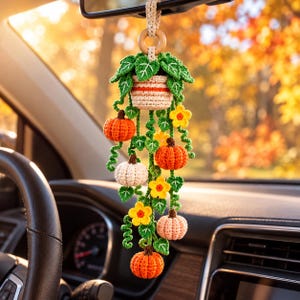 Könnte beinhalten: Ein gehäkelter Rückspiegelanhänger für das Auto, bestehend aus einer kleinen Topfpflanze mit grünen Blättern, orangefarbenen und weißen Kürbissen sowie gelben Blumen, befestigt an einem glatten Holzring.