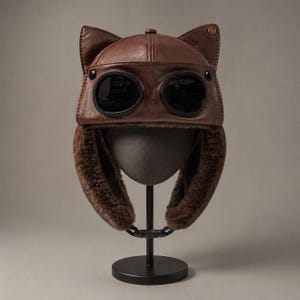 May include: A brown leather aviator-style hat featuring two pointed cat-like ears on the crown, integrated dark-tinted circular goggles on the front, and thick brown faux fur lining on the ear flaps, which are secured by a black plastic buckle.