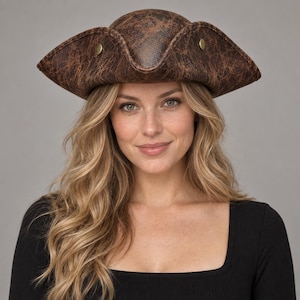 Peut inclure: Un chapeau tricorne marron présentant une texture vieillie et usée avec un aspect tacheté. Le chapeau possède un large bord relevé sur trois côtés et est orné de deux petits boutons circulaires métalliques de couleur laiton sur les côtés.