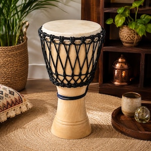 May include: A djembe drum featuring a light-colored wooden body with a smooth finish and a circular drumhead. The top section is secured with a black rope lacing system arranged in a diamond pattern, and a dark cord is wrapped around the narrow base.