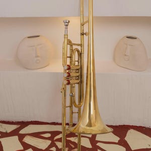 Op de afbeelding: Een koperen ventieltrombone met een lange, slanke beker, drie verticale ventielen met rode accenten en een zilverkleurig mondstuk. Het instrument is vervaardigd uit gepolijst geel koper met een gebogen schuif en verstevigingen.
