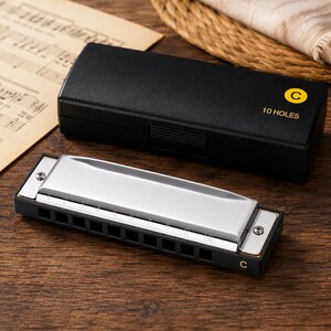 May include: A silver-coloured harmonica with ten numbered holes and a black body, accompanied by a black rectangular storage case featuring the text C and 10 HOLES printed in yellow on the top lid. The instrument is made of metal and plastic.