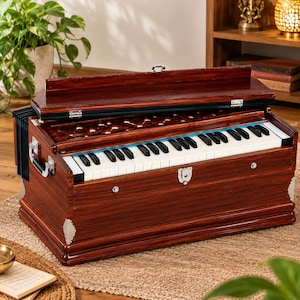 May include: A dark brown wooden harmonium featuring a keyboard with white and black keys, a bellows system on the side, and a decorative carved wooden panel above the keys. The instrument includes silver-toned metal latches and corner accents.