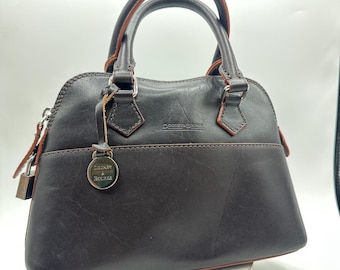 Peut inclure: Un sac à main en cuir marron foncé avec deux poignées supérieures et une breloque en métal argenté suspendue à la base d'une poignée. Le sac présente une fermeture éclair, une couture horizontale et le logo Dooney & Bourke gravé.