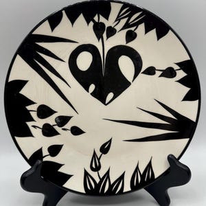 May include: A round ceramic plate featuring a high-contrast black and white design. The surface displays stylized botanical motifs, including large heart-shaped leaves, pointed foliage, and small dangling leaf clusters arranged in a circular pattern.