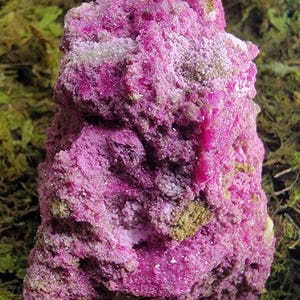 May include: A raw, crystalline mineral specimen featuring a vibrant magenta and deep pink coloration. The surface is highly textured with jagged, uneven edges and small, sparkling crystal formations throughout the dense, rocky structure.