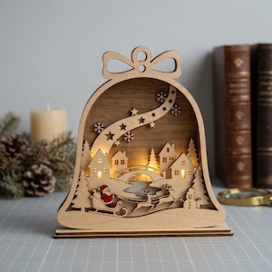 May include: A bell-shaped wooden light display featuring a laser-cut winter village scene with houses, a bridge, a snowman, and Santa Claus in a sleigh pulled by reindeer. The scene is illuminated from within and decorated with stars and snowflakes.