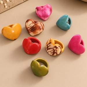May include: A collection of eight heart-shaped rings in various colors including solid yellow, red, olive green, bright pink, and teal, along with two marbled patterns in brown and white. The text Casa Fátima Ecuadorian Artisan Collection is visible.