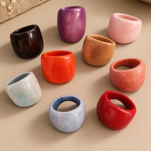 May include: A collection of nine chunky, rounded rings in various solid and marbled colors including dark brown, purple, light pink, orange, tan, coral, light blue, and red. The text "CASA Fátima ECUADORIAN ARTISAN COLLECTION" appears at the bottom.