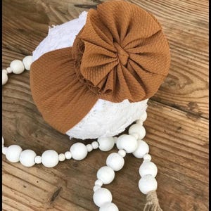 Puede incluir: Una diadema de tela texturizada color marrón mostaza con un gran detalle de roseta circular fruncida, colocada sobre una esfera cubierta de encaje blanco, acompañada por una larga hilera de cuentas de madera blancas, lisas y redondas, con una pequeña borla de yute.