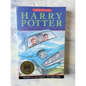 May include: A paperback book titled Harry Potter and the Chamber of Secrets by J.K. Rowling. The cover features a light blue flying car with two people and an owl inside. A gold Smarties Book Prize sticker is on the lower left corner.