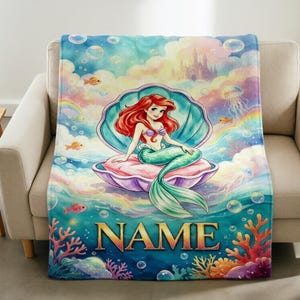 Puede incluir: Una manta suave con una vibrante ilustración en acuarela de una sirena de pelo largo y rojo sentada dentro de una gran concha abierta. El fondo muestra una escena submarina con corales coloridos, burbujas, medusas y un castillo.