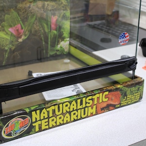 Op de afbeelding: Een Zoo Med Naturalistic Terrarium met een glazen behuizing, zwart frame en een bedrukte kartonnen voet met een reptielafbeelding. Het glas toont een achtergrond van tropische planten en een kleine ronde sticker met de Amerikaanse vlag.
