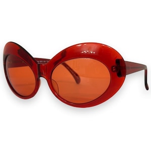 Könnte beinhalten: Übergroße Sonnenbrille mit einem transparenten roten Kunststoffrahmen und passenden rot getönten Gläsern. Die Fassung hat eine markante, breite und abgerundete Form mit dicken Rändern und integrierten Bügeln an den äußeren Seiten der Gläser.