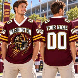 May include: A burgundy football jersey with white and gold accents, featuring a V-neck collar and short sleeves. The front displays the text WASHINGTON above a graphic of a gladiator helmet. The back shows the text YOUR NAME and the number 00.