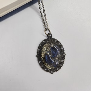 以下が含まれることがあります： 青い金色の楕円形のペンダントが付いた金のチェーンネックレス。ペンダントは装飾的なフィリグリーフレームと、青と金のグリッターが入ったレジンインレイが付いています。