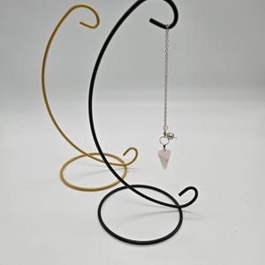 May include: A gold and black metal pendulum stand with a rose quartz crystal pendulum. The stand has a circular base and a curved design. The pendulum hangs from a chain.