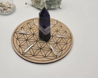 May include: A dark purple and white crystal tower sits on a wooden coaster with a black geometric pattern. Clear quartz crystals are arranged around the tower. A green rock and a glass container with small white objects are in the background.