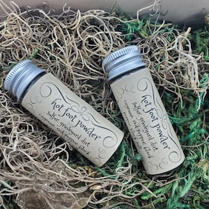 May include: Two small glass bottles with silver lids, labeled "hot foot powder." The labels list ingredients like sulfur, graveyard dirt, and cayenne pepper. The bottles rest in a bed of dried moss and twigs.