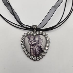 Könnte beinhalten: Eine schwarze Bandkette mit einem herzförmigen Anhänger. Der Anhänger ist mit Strasssteinen verziert und zeigt eine Schwarz-Weiß-Anatomieillustration.