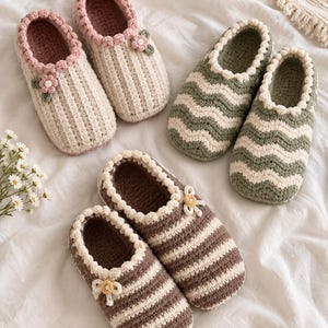 Op de afbeelding: Drie paar handgemaakte gehaakte slippers. Eén paar is crème met een roze rand en kleine bloemaccenten. Een ander paar heeft een groen en crème golvend patroon. Het derde paar is bruin en crème gestreept met kleine gele bloemaccenten.