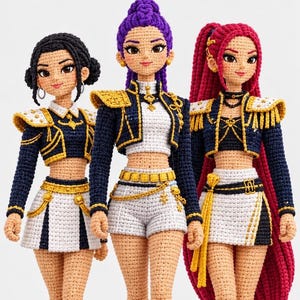 Pode incluir: Três bonecas de crochê feitas à mão com pele bronzeada e roupas detalhadas. Cada boneca usa uma jaqueta curta azul-marinho com detalhes dourados, um top branco e uma saia ou shorts brancos. Têm penteados feitos de fios pretos, roxos e vermelhos.