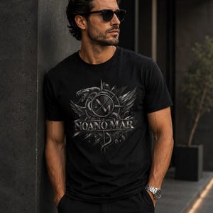 May include: A black short-sleeved t-shirt featuring a graphic print on the chest. The design includes a stylised snake, a compass-like emblem with the letters NM, and the text NOANO MAR in a metallic silver font against a dark background.