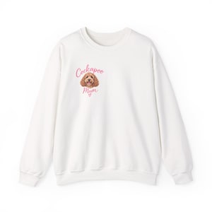 Puede incluir: Una sudadera blanca de manga larga con un gráfico en la parte superior izquierda del pecho. El gráfico muestra una ilustración de un perro Cockapoo marrón con el texto rosa "Cockapoo Mum" escrito encima y debajo, con un pequeño corazón rosa.