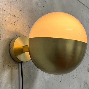 Könnte beinhalten: Eine Wandleuchte mit einem kugelförmigen Design, bestehend aus einem Sockel aus gebürstetem Messing und einer Kugel aus transluzentem weißem Glas. Die Lampe strahlt ein warmes Licht aus der oberen Hälfte ab, mit einem dünnen schwarzen Stromkabel.
