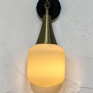 Könnte beinhalten: Eine Wandleuchte mit einem zylindrischen, mattweißen Glasschirm, der an einem konischen, messingfarben gebürsteten Metallhals befestigt ist. Die Leuchte ist mit einer runden, mattschwarzen Montageplatte mit Metallöse an der Wand befestigt.