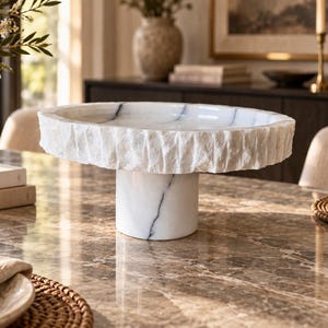 May include: A white marble pedestal bowl featuring a smooth, polished interior surface with grey veining and a contrasting rough, chiseled exterior edge. The bowl is supported by a cylindrical marble base that matches the white and grey veining.