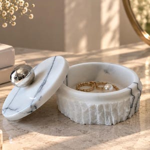 May include: A round white marble jewellery box with grey veining and a textured, rough-hewn exterior base. The lid features a smooth, polished surface with a central, rounded, metallic silver handle. Inside the open container sits a gold ring.