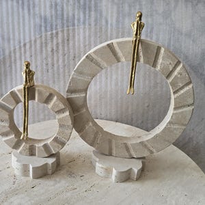 Pu&ograve; includere: Due sculture circolari in pietra travertino beige, ciascuna con una piccola figura in metallo dorato seduta sul bordo superiore. Gli anelli in pietra presentano una superficie testurizzata e scanalata, montati su basi in pietra di diverse dimensioni.