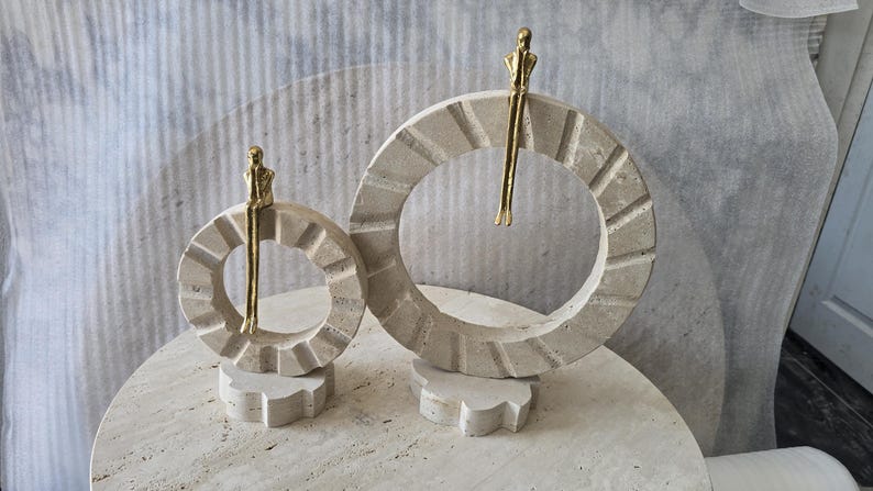 Pu&ograve; includere: Due sculture circolari in pietra travertino beige, ciascuna con una piccola figura in metallo dorato seduta sul bordo superiore. Gli anelli in pietra presentano una superficie testurizzata e scanalata, montati su basi in pietra di diverse dimensioni.