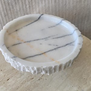 May include: A circular white marble tray featuring prominent dark gray veining and subtle orange streaks. The interior surface is smooth and polished, while the exterior rim displays a rough, chiseled texture with vertical grooves around the edge.