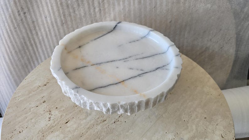 May include: A circular white marble tray featuring prominent dark gray veining and subtle orange streaks. The interior surface is smooth and polished, while the exterior rim displays a rough, chiseled texture with vertical grooves around the edge.