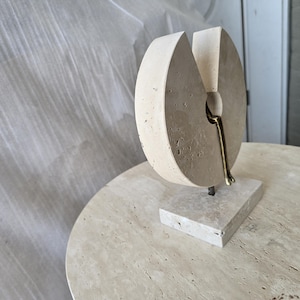 K&ouml;nnte beinhalten: Eine beige Steinskulptur mit kreisf&ouml;rmiger Form und keilf&ouml;rmigem Ausschnitt. Eine kleine, metallische, bronzefarbene Figur ist in der &Ouml;ffnung positioniert und scheint auf dem Rand zu sitzen. Die Skulptur ruht auf einem rechteckigen Steinsockel.