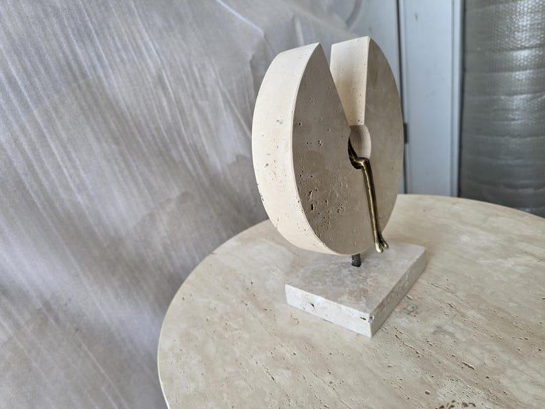 K&ouml;nnte beinhalten: Eine beige Steinskulptur mit kreisf&ouml;rmiger Form und keilf&ouml;rmigem Ausschnitt. Eine kleine, metallische, bronzefarbene Figur ist in der &Ouml;ffnung positioniert und scheint auf dem Rand zu sitzen. Die Skulptur ruht auf einem rechteckigen Steinsockel.