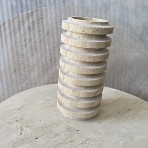 May include: A cylindrical vase crafted from beige travertine stone, featuring a series of stacked, horizontal circular discs that create a ribbed, textured exterior surface with a natural porous finish and a hollow opening at the top.