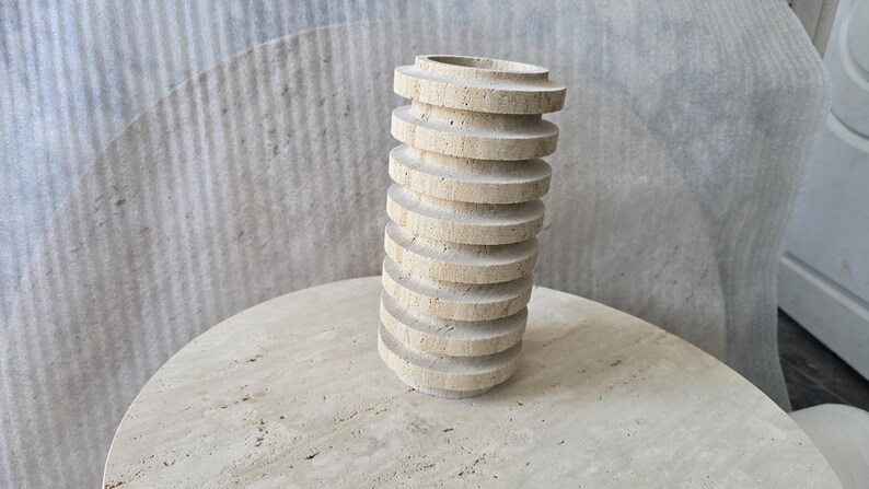 May include: A cylindrical vase crafted from beige travertine stone, featuring a series of stacked, horizontal circular discs that create a ribbed, textured exterior surface with a natural porous finish and a hollow opening at the top.