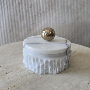 May include: A round white marble container with grey veining, featuring a textured, rough-hewn exterior surface and a smooth, flat lid. A polished, metallic gold-toned spherical knob with a dimpled, hammered texture sits atop the centre of the lid.