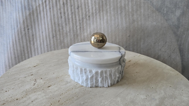 May include: A round white marble container with grey veining, featuring a textured, rough-hewn exterior surface and a smooth, flat lid. A polished, metallic gold-toned spherical knob with a dimpled, hammered texture sits atop the centre of the lid.